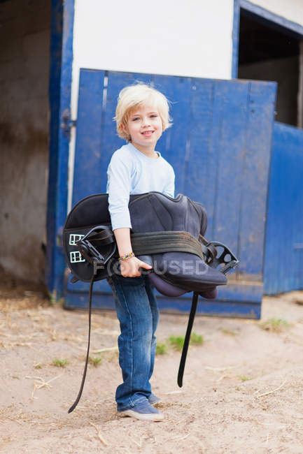 Boy carrying horse saddle, focus on foreground — Stock Photo
