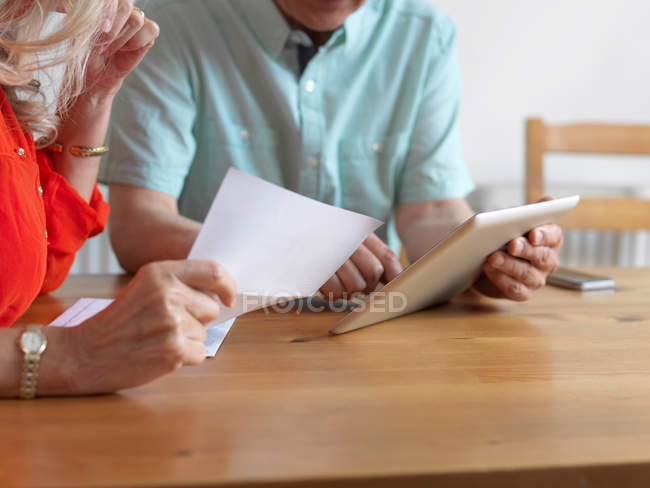 Pareja mayor leyendo correo en la cocina, sosteniendo hoja de papel y usando tableta digital - foto de stock