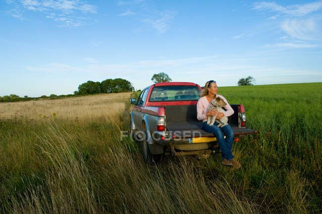 Mulher sentada na parte de trás de pegar caminhão em um campo, segurando um cão — Fotografia de Stock