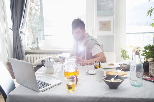 Молодий чоловік снідає і користується ноутбуком. — стокове фото