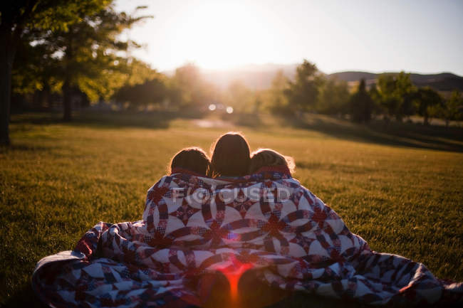 Vista trasera de tres niñas envueltas en manta al atardecer - foto de stock