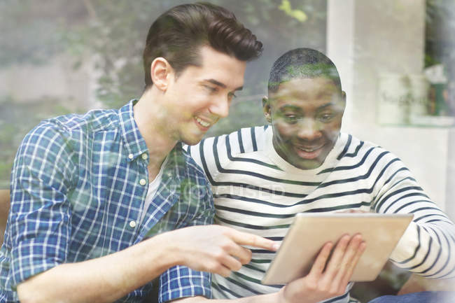 Zwei junge männliche Freunde blicken hinter Fenster auf digitales Tablet — Stockfoto