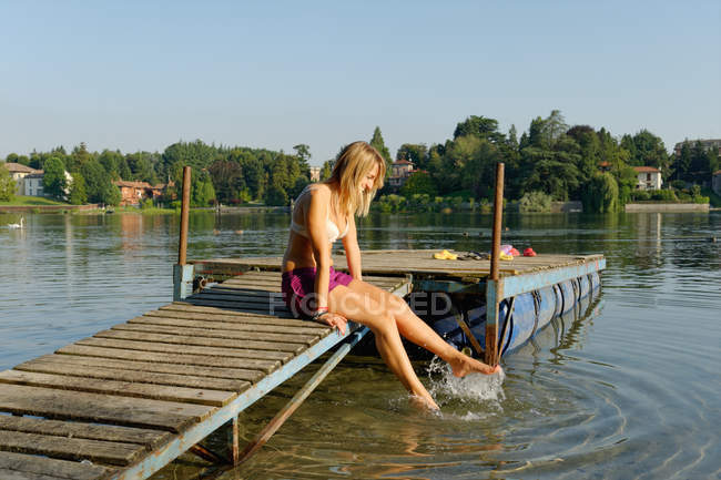 Teilweise bekleidete erwachsene Frau, auf Steg sitzend, Füße im Wasser tauchend — Stockfoto