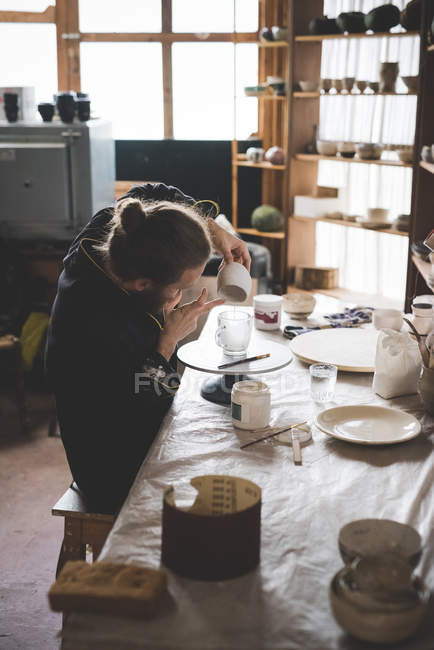Seitenansicht eines erwachsenen Mannes, der in der Werkstatt sitzt und Keramikglasur auf Tontopf aufträgt — Stockfoto