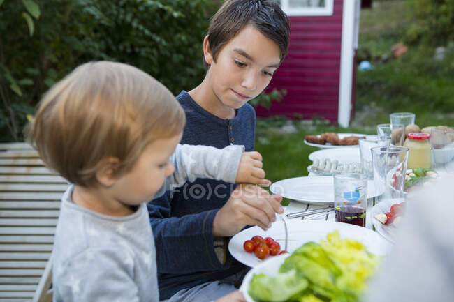 Teenage boy helping female toddler eat food at garden barbecue — Stock Photo