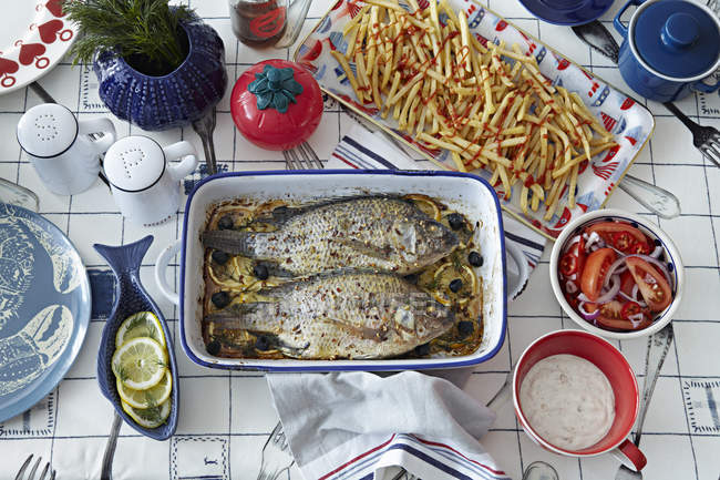 Запечённая рыба на столе с картошкой фри и приправами — стоковое фото
