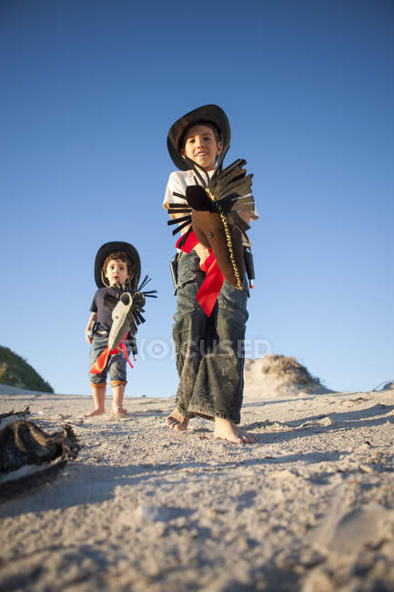 Low angle view of two brothers dressed as cowboys with hobby horses in sand dunes — Stock Photo