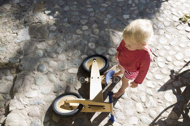 Overhead view of female toddler and wooden bicycle on cobbles — Stock Photo