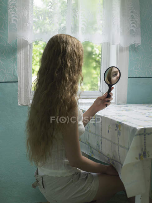 Отражение молодой женщины, смотрящей в ручное зеркало на кухне — стоковое фото