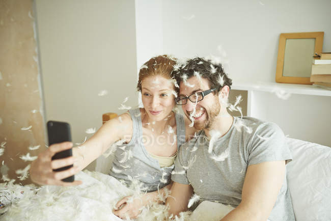 Casal adulto médio coberto de penas de luta de travesseiro levando selfie smartphone na cama — Fotografia de Stock