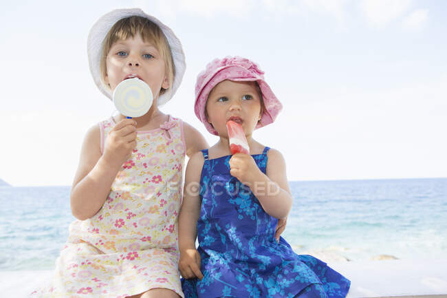 Niña y hermana usando sombreros comiendo lollies de hielo en la playa - foto de stock