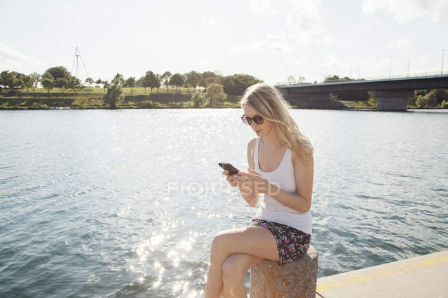 Mujer joven sentada a la orilla del río mensajes de texto en el teléfono inteligente, Isla del Danubio, Viena, Austria - foto de stock