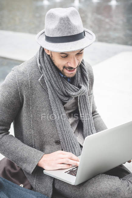 Улыбающийся бизнесмен сидит на стене и пользуется ноутбуком — стоковое фото