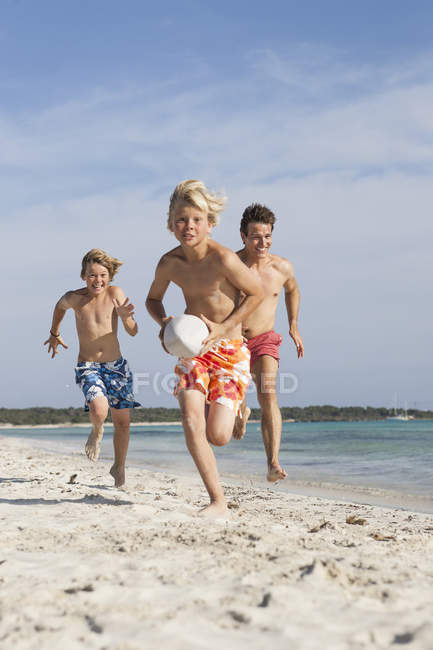 Menino correndo com bola de rugby perseguido por irmão e pai na praia, Maiorca, Espanha — Fotografia de Stock