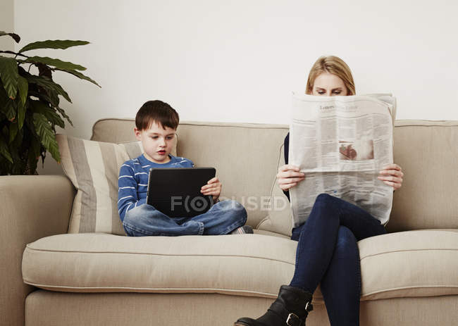 Junge nutzt digitales Tablet, Mutter liest Zeitung — Stockfoto