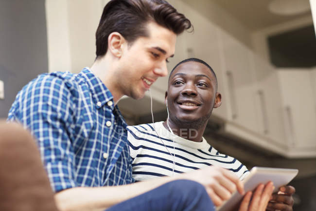Два молодых человека слушают музыку на цифровом планшете на диване в гостиной — стоковое фото