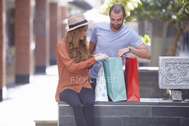 Jovem mulher sentada na parede com sacos de compras, homem olhando em sacos — Fotografia de Stock