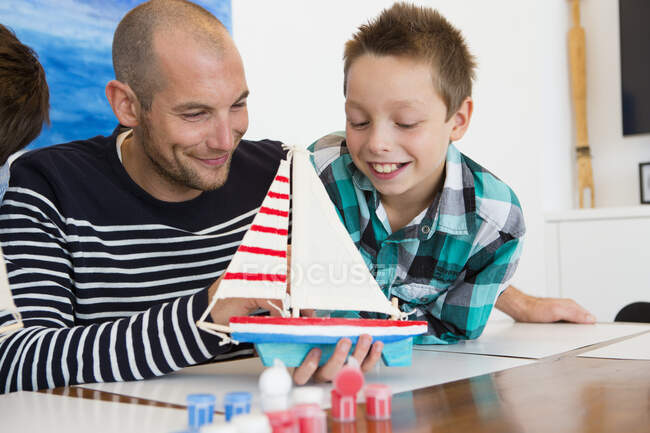 Homem adulto médio e filho admirando barco de brinquedo pintado na mesa da cozinha — Fotografia de Stock