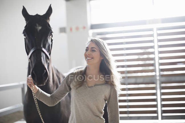 Young woman leading black horse in stables — Stock Photo
