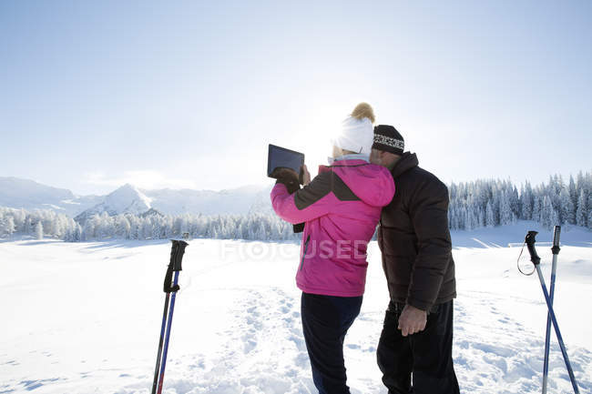 Vista trasera de la pareja de personas mayores en el paisaje nevado utilizando tableta digital para tomar una foto de la cordillera, Sattelbergalm, Tirol, Austria - foto de stock