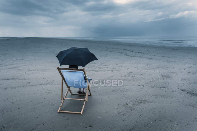 Mujer madura sentada en la silla de cubierta en la playa tormentosa, bajo el paraguas - foto de stock