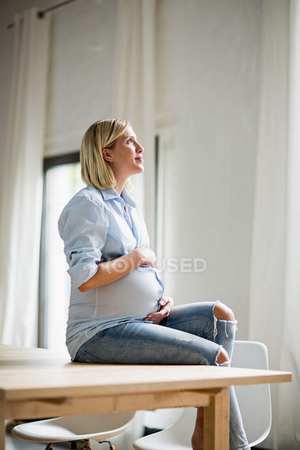 Gravidanza completa giovane donna seduta a tavola con lo stomaco in mano — Foto stock
