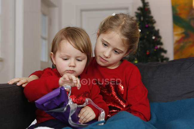 Sisters sitting together on sofa looking down opening gift — Stock Photo