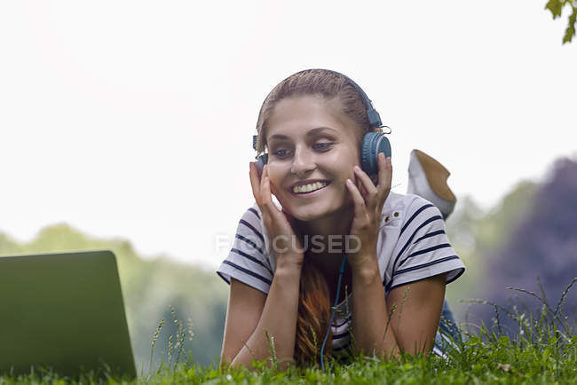 Молода жінка в навушниках лежить на траві спить на ліктях, дивлячись на ноутбук посміхаючись — стокове фото