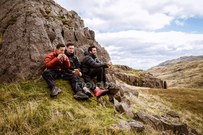 Three young male hiking friends eating sandwiches, The Lake District, Cumbria, UK — Stock Photo