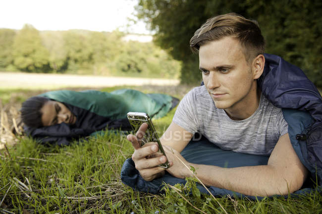 Young man lying sleeping bag texting on smartphone in field — Stock Photo