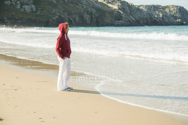 Mulher madura de pé na praia com as mãos nos bolsos, Camaret-sur-mer, Bretanha, França — Fotografia de Stock