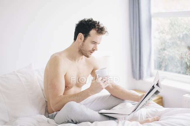 Junger Mann liegt im Bett, trinkt Kaffee und liest Zeitung — Stockfoto