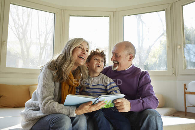 Бабушка с дедушкой у окна читают книгу с внуком, улыбаются — стоковое фото