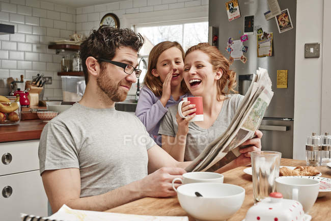 Средний взрослый мужчина за завтраком читает газету, пока дочь и мать смеются — стоковое фото