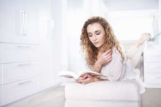 Mujer joven en otomana leyendo un libro en el dormitorio - foto de stock