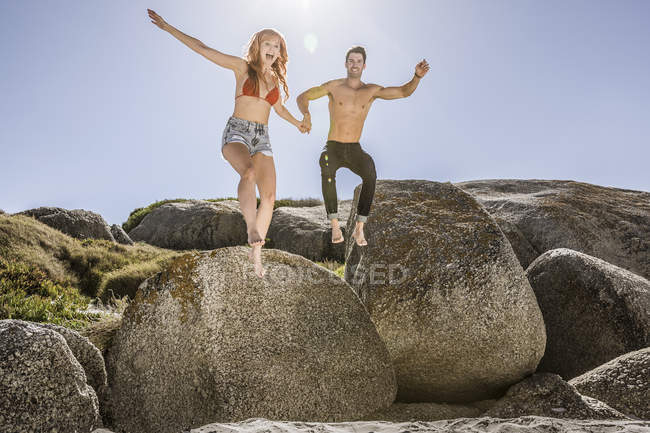 Couple outdoors, holding hands, jumping off of rock onto sand — Stock Photo