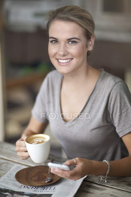 Портрет молодой женщины, сидящей в кафе с чашкой кофе и сотовым телефоном — стоковое фото