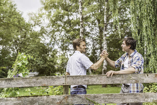 Two male adult friends shaking hands at garden fence — Stock Photo