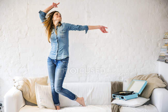 Молодая женщина, стоящая на диване, танцуя под винтажный проигрыватель — стоковое фото