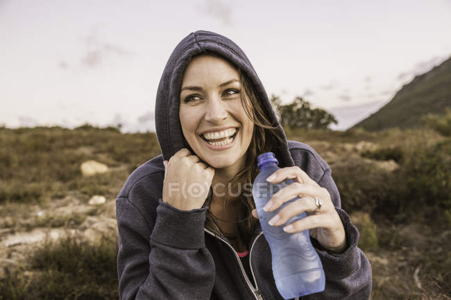 Повнометражний вид спереду жінки в капюшоні зверху тримає пляшку з водою, дивиться геть усміхнений — стокове фото