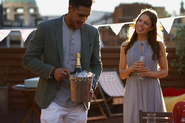 Casal adulto médio na festa no terraço do telhado segurando balde de gelo com champanhe sorrindo — Fotografia de Stock