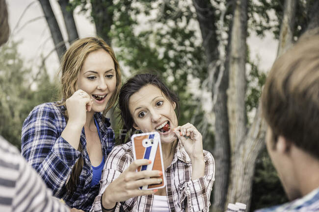 Vue d'épaule de jeunes femmes mangeant des cerises prenant selfie smartphone — Photo de stock