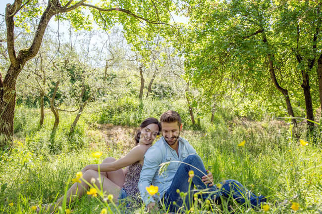 Junges Paar sitzt Rücken an Rücken auf dem Gras und blickt lächelnd nach unten — Stockfoto