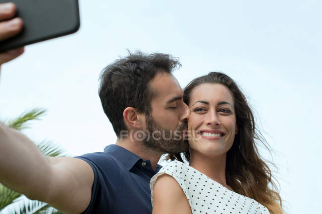 Молодий чоловік цілує дівчину на щоці для смартфона селфі — стокове фото