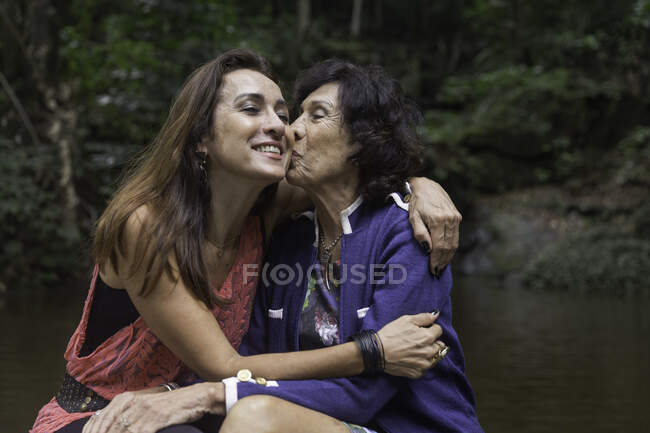 Старша жінка цілує дочку на щоці на лісовій річці — стокове фото