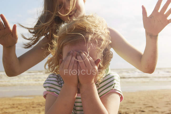 Chica cubriendo los ojos delante de su hermana en la playa, Camber Sands, Kent, Reino Unido - foto de stock