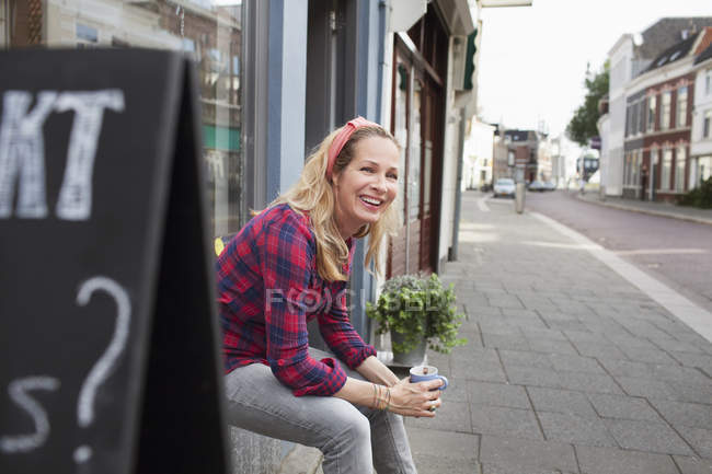 Mulher sentada na janela da loja segurando xícara de café olhando para longe sorrindo — Fotografia de Stock