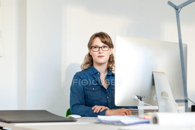 Mujer joven con anteojos sentada en el escritorio usando la computadora mirando hacia otro lado sonriendo - foto de stock