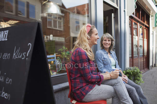 Mujeres sentadas en sillas frente a la tienda sosteniendo tazas de café sonriendo - foto de stock