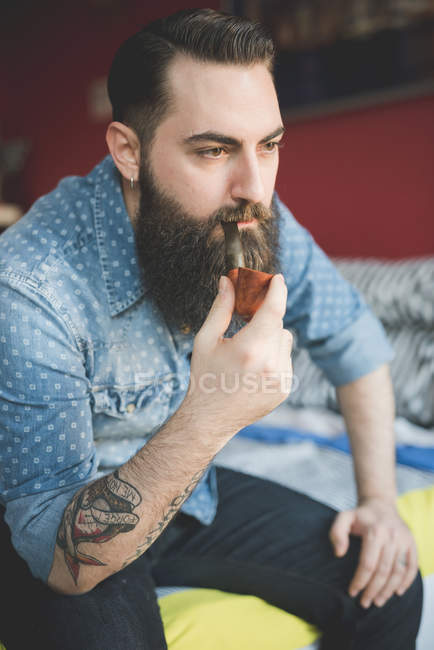 Молодий бородатий чоловік палить трубу на ліжку — стокове фото
